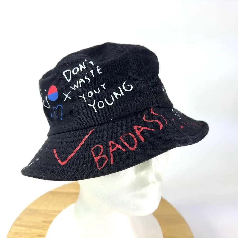 Black cotton bucket hat with handwritten punk slogans and red graffiti, front-facing view showing 'Don't Waste Your Young' te