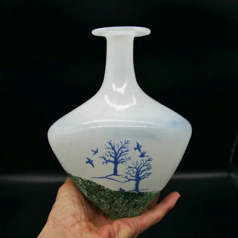 Kosta Boda October vase by Kjell Engman, white hand-blown glass with blue tree and bird silhouettes, held in palm showing tex