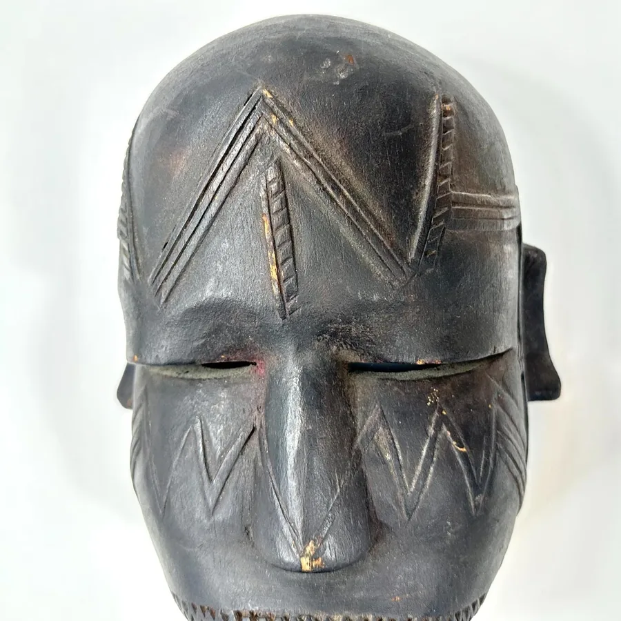 Angled mask view displaying forehead geometric carvings, nose projection, ear detail, striped mouth, subtle surface weatherin