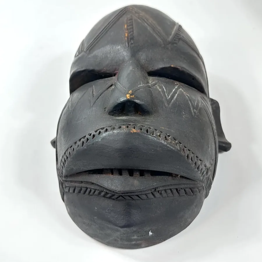 Close frontal view showing striped mouth carving, eye slit aperture, aged dark patina, geometric facial patterns clearly visi