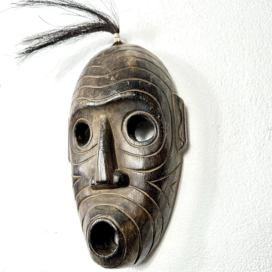 Hand-carved wooden African mask with geometric ridges, horsehair tuft, and expressive face mounted on white wall