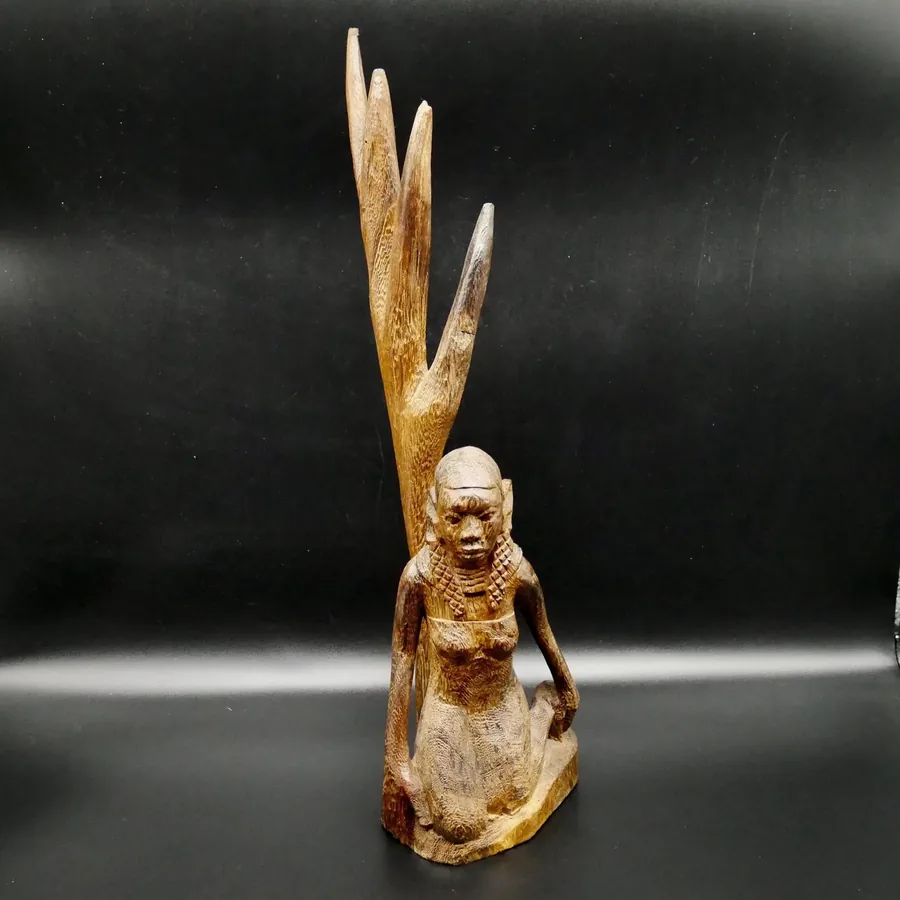 Hand-carved wooden tribal woman sculpture with natural wood grain patina, seated beside tall textured tree branch, 40cm folk