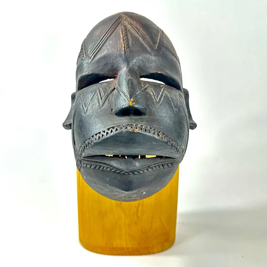 Mounted mask on wood stand showing dark wood carving with geometric patterns, museum-style presentation with supporting base