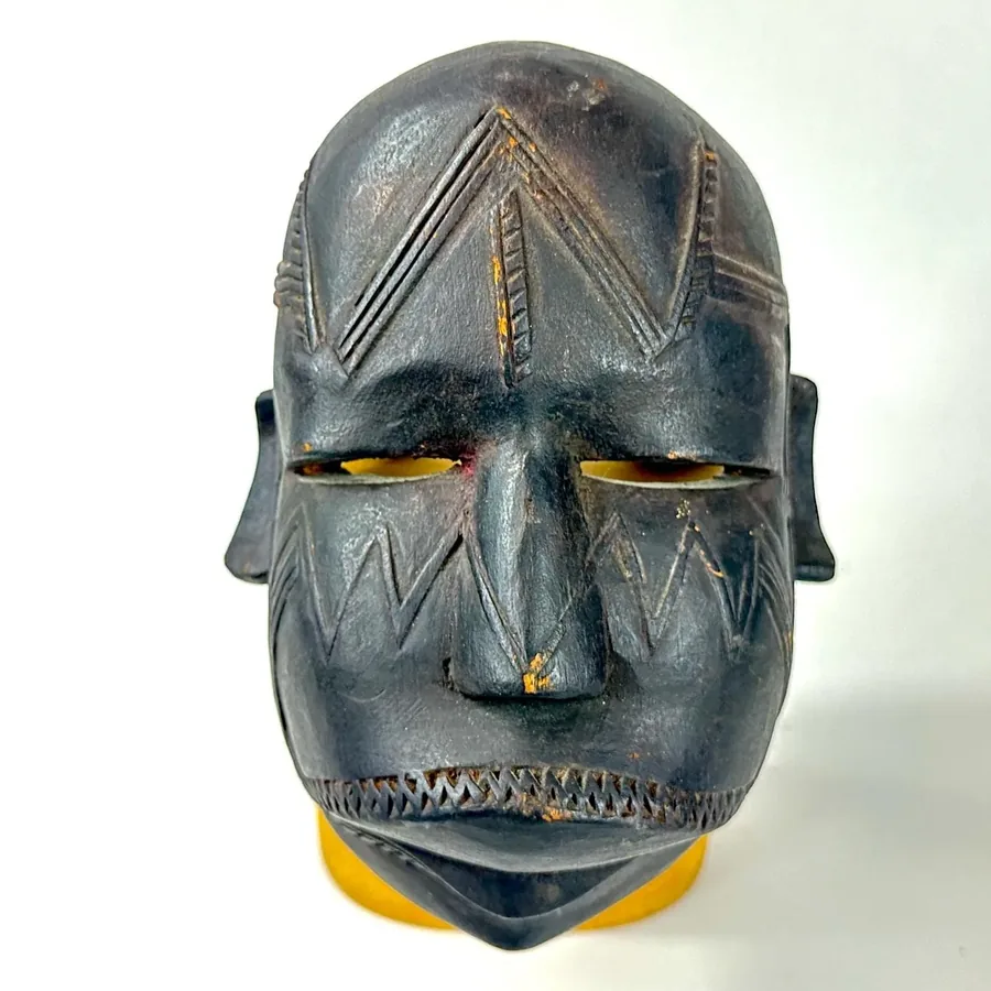 Mounted vintage tribal mask on golden-yellow wood base, dramatic lighting on carved features, geometric patterns emphasized,