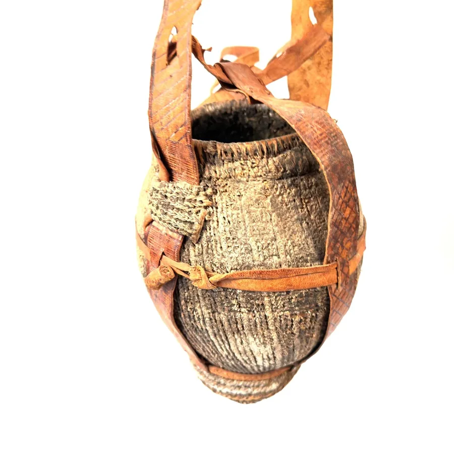 Vintage tribal water vessel with woven reed base, mud coating, dual leather shoulder straps, front view