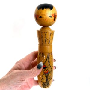 Vintage Japanese kokeshi doll held in hand, golden wood with black-painted head, hand-painted kimono scene and calligraphy de