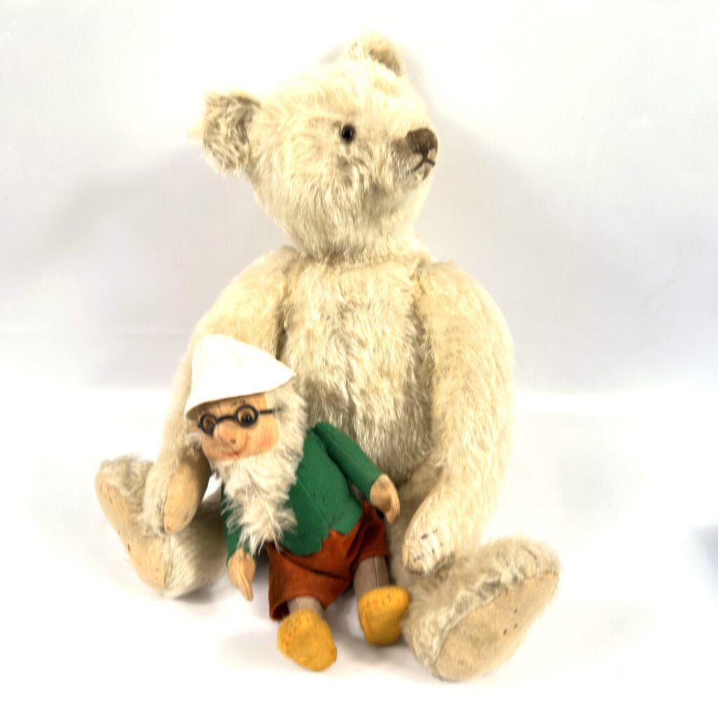 Cream mohair teddy bear seated beside a small jointed doll wearing green and red clothing, vintage companion pair portrait