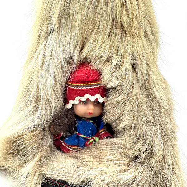 Close-up of doll's face wearing red woolen hat with white embroidered trim and blue fabric tunic with decorative details