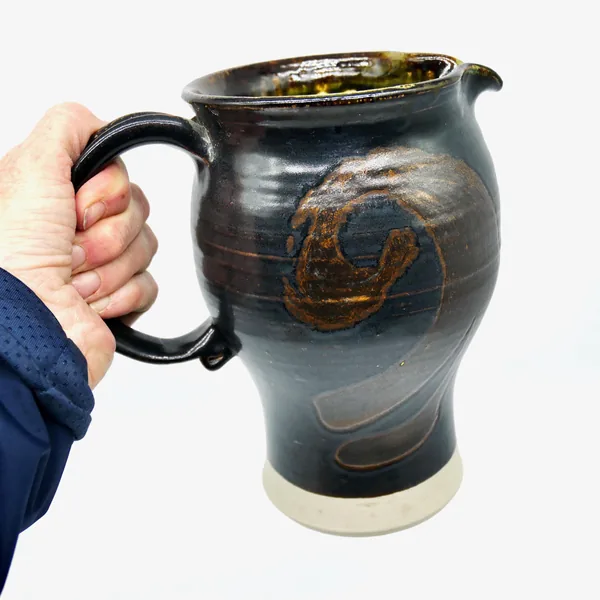 Hand holding dark brown stoneware pitcher, showing scale, texture of glaze, handle ergonomics, and realistic size perspective
