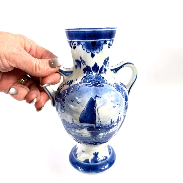 Hand holding Delft blue vase by handle, revealing side profile with sailing ship and floral border details