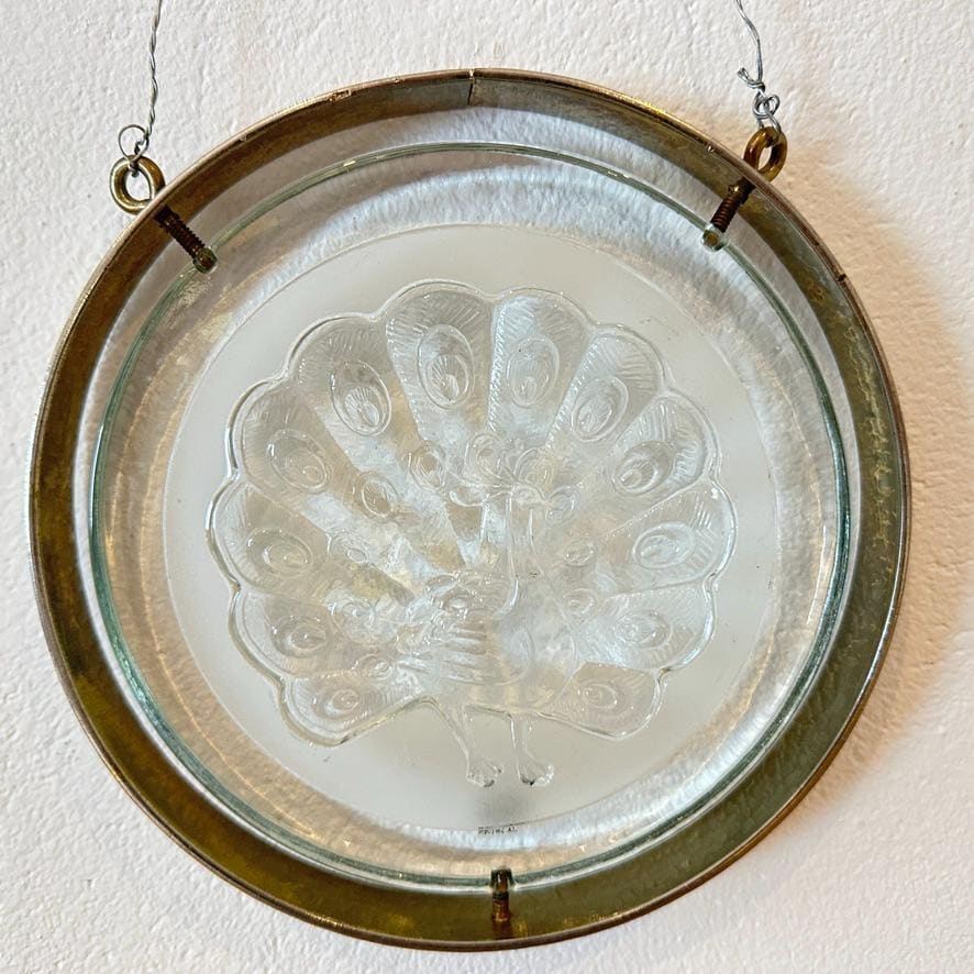 Overhead view of suspended peacock glass wall plaque showing wire hangers, brass frame, and complete embossed design