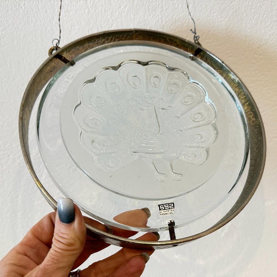 Peacock glass plaque in hand angled to show glass dimension, brass frame patina, and how light interacts with embossed relief