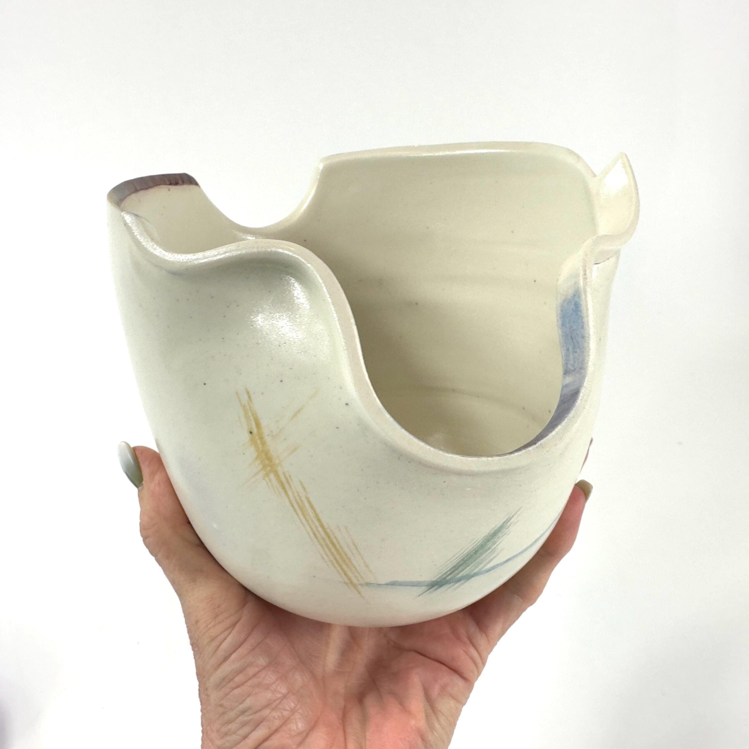Pottery bowl cradled in two hands displaying full sculptural form, cream body with layered abstract glaze in blue and olive t