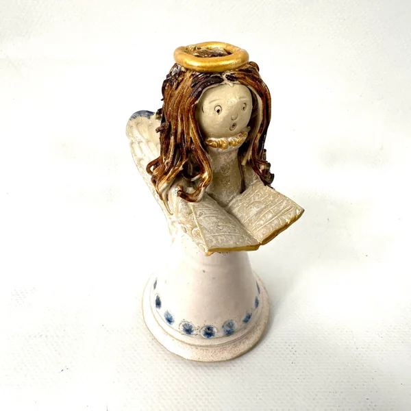 Side profile of ceramic angel showing textured spaghetti hair, patterned wings, and songbook held at chest, against neutral b