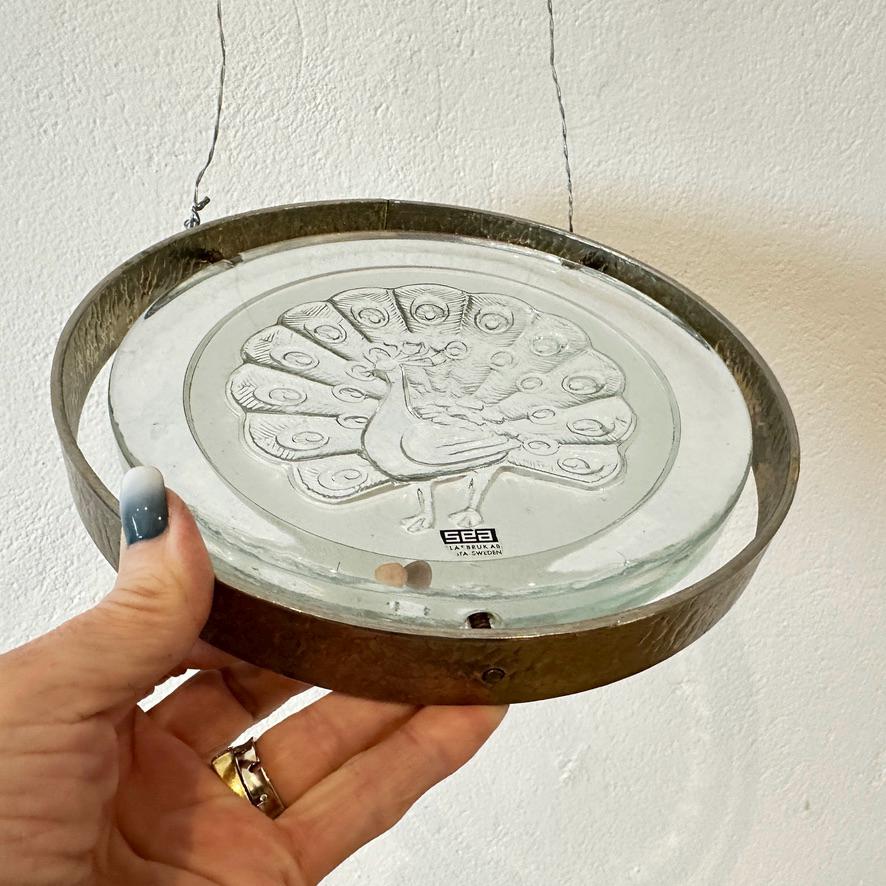 Swedish peacock glass plaque centered in hands showing complete circular form, metal mounting frame all visible from front