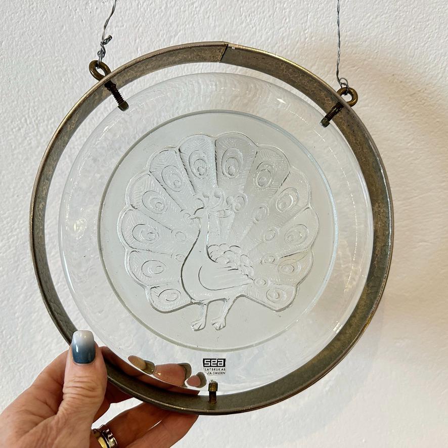Three-quarter angle of glass plaque in hand showing peacock design, glass depth, and original brass frame together