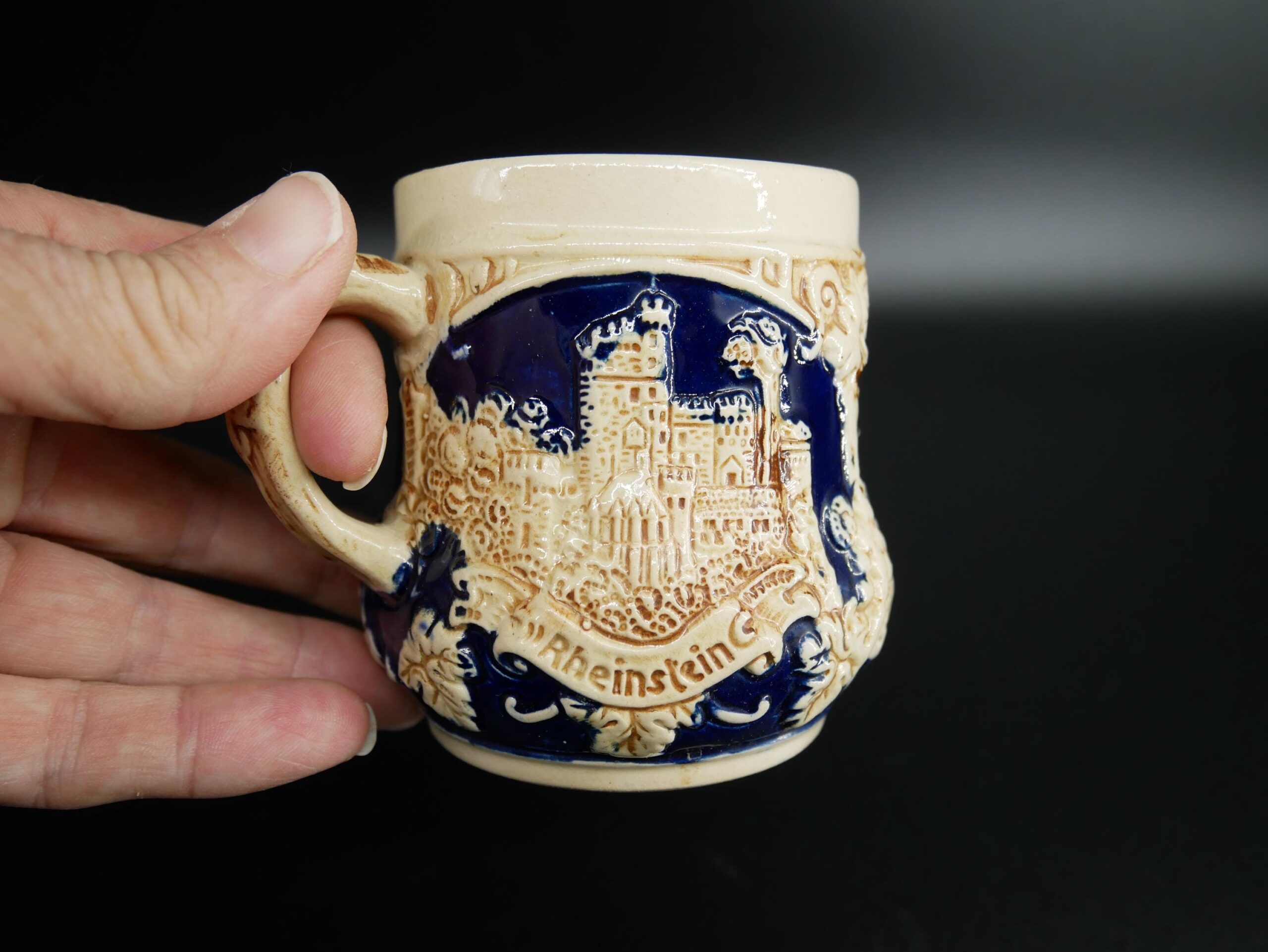 Vintage West Germany ceramic beer stein in cream and cobalt blue with embossed castle imagery and ornate gilt detailing, held