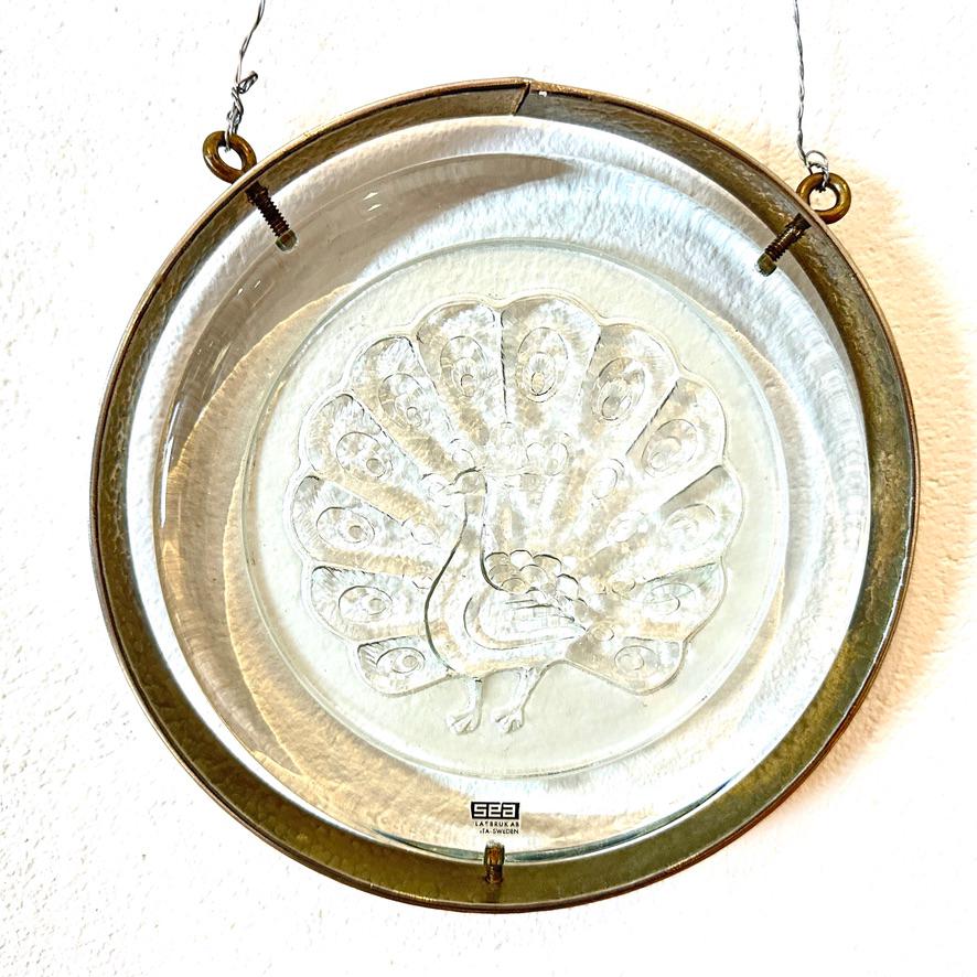 Wall-mounted peacock glass plaque with patinated brass frame, flat lay view showing full circular design and metal hardware