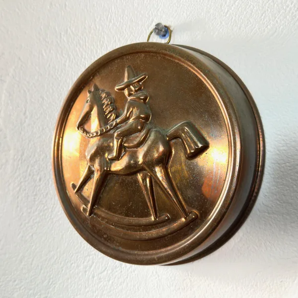 Warm-toned photograph of copper jelly mold with folk art relief, brass loop and tin interior visible, warm lighting