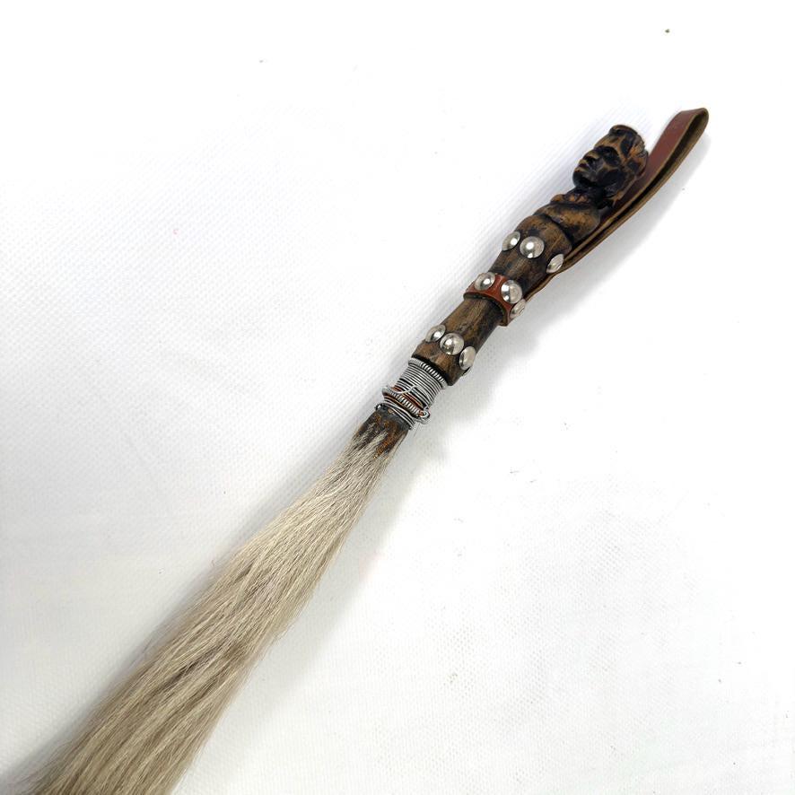 Close-up of carved wooden handle showing stylised head motif, metal studwork, and leather bands, full whisk length visible