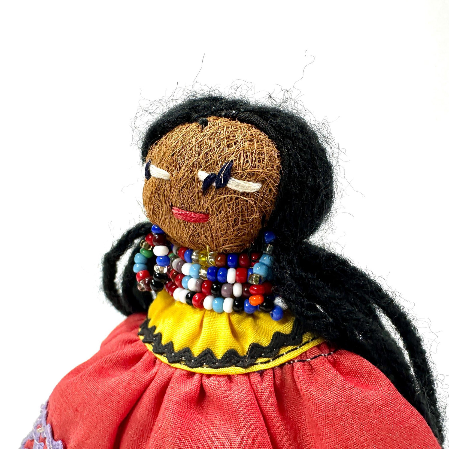 Close-up portrait of doll's face and upper body with corn-husk features, beaded necklace, and yellow ribbon trim accent