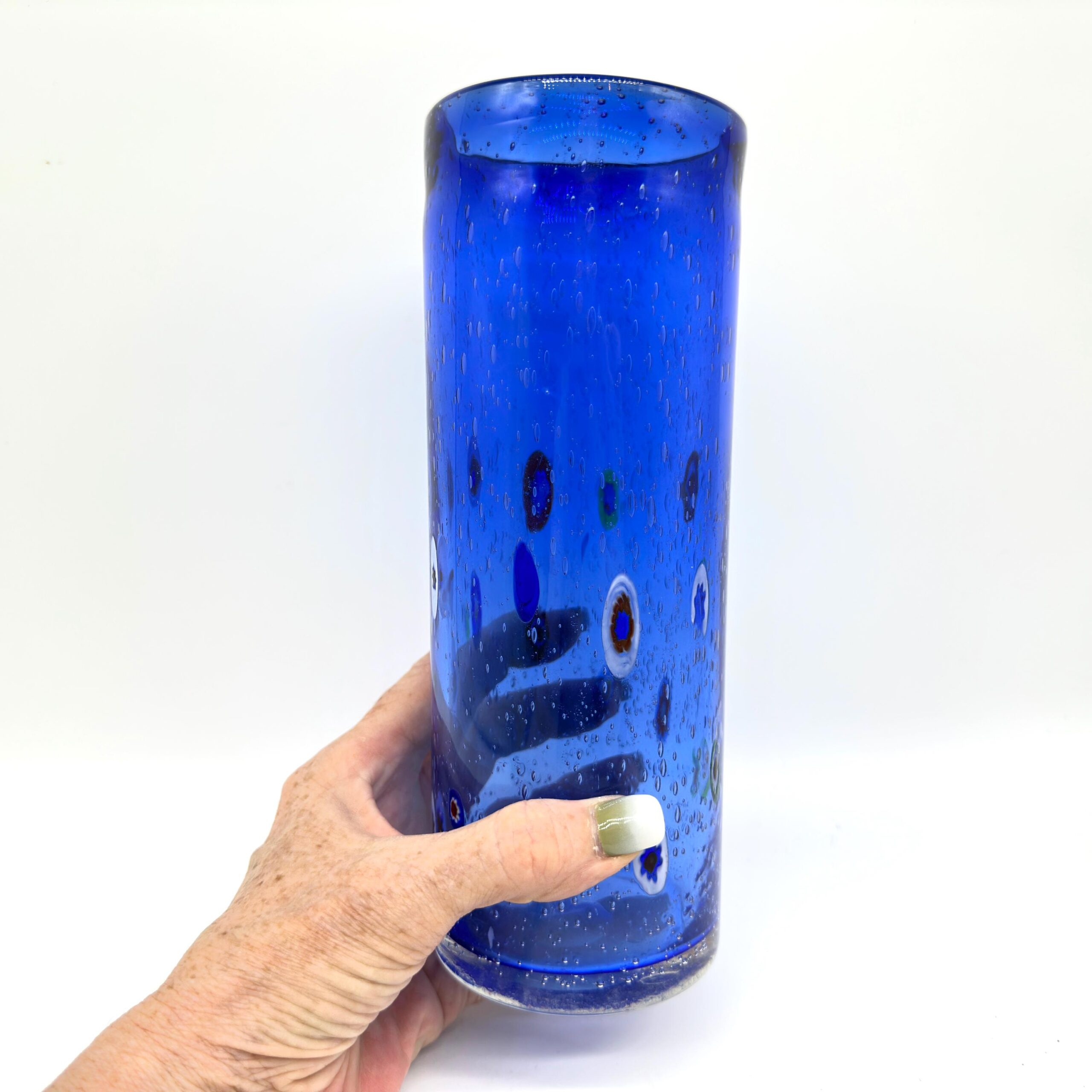 Deep cobalt blue Murano glass cylinder vase with colorful millefiori canes and bubble inclusions, held in hand to show scale