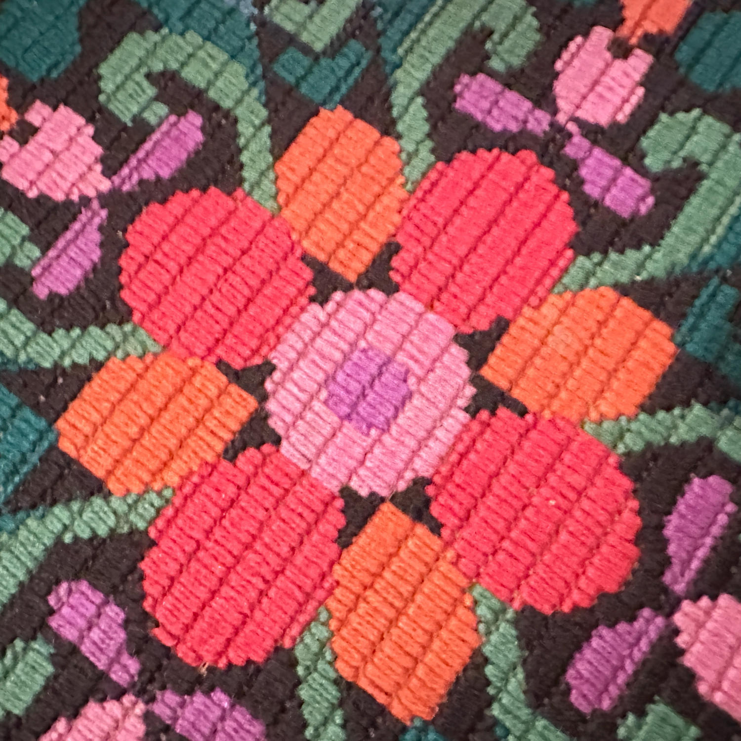 Extreme close-up of hand-stitched needlepoint flowers, individual cross-stitches visible, coral pink orange lavender blooms,