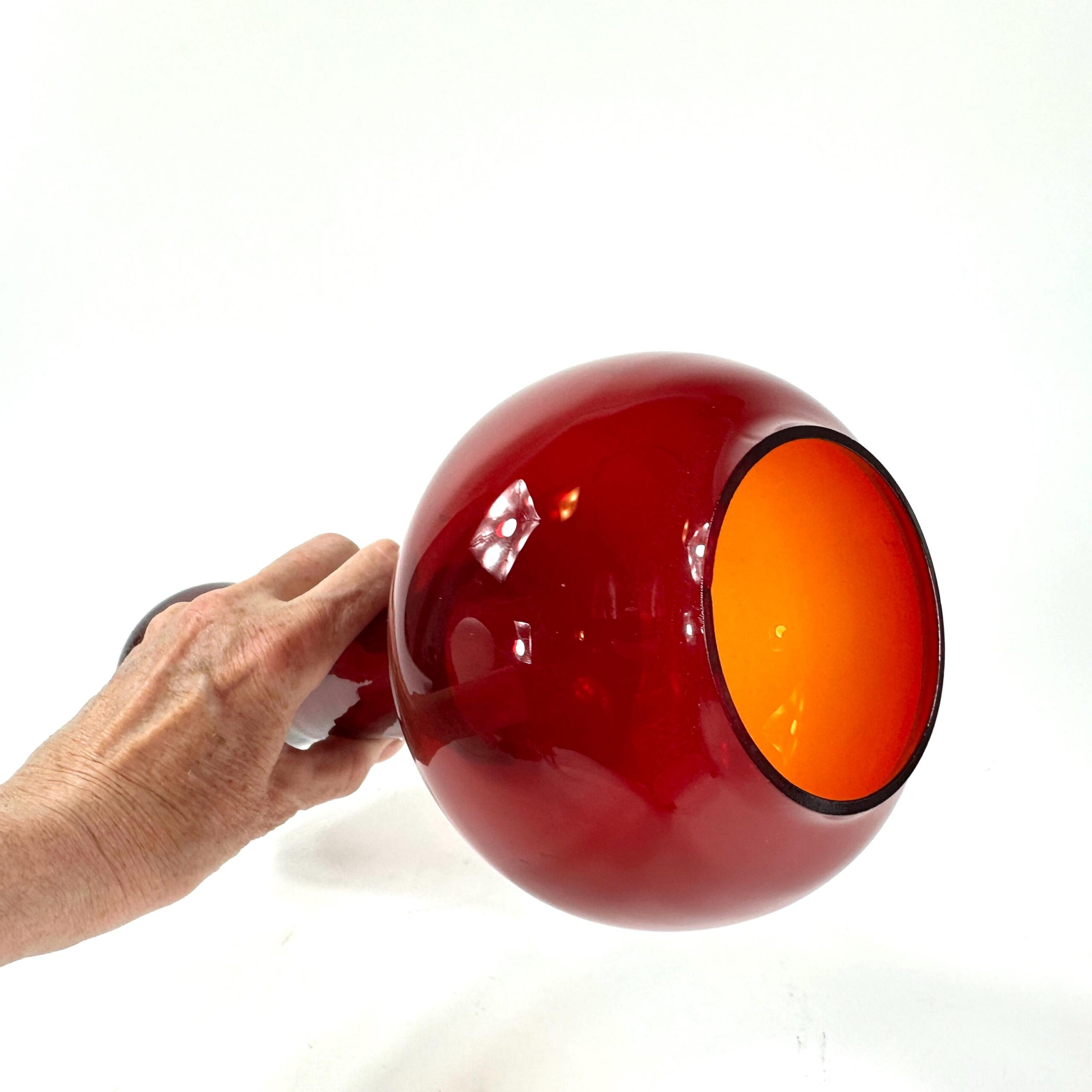 Hand holding Åseda vase revealing warm orange interior glow of cased glass, ruby red exterior under natural light
