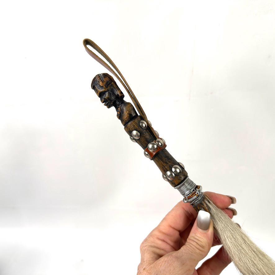 Hand holding fly whisk grip showing carved head detail, leather bands, and metal work up close with horsehair extending