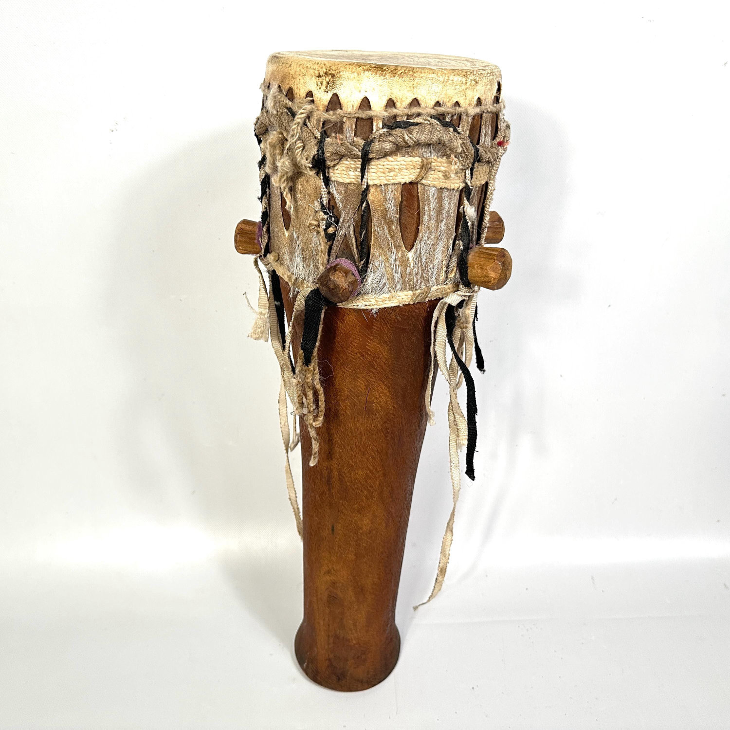 Single large African hand drum with natural hide head, carved wood body, and dangling rope tuning system photographed in prof