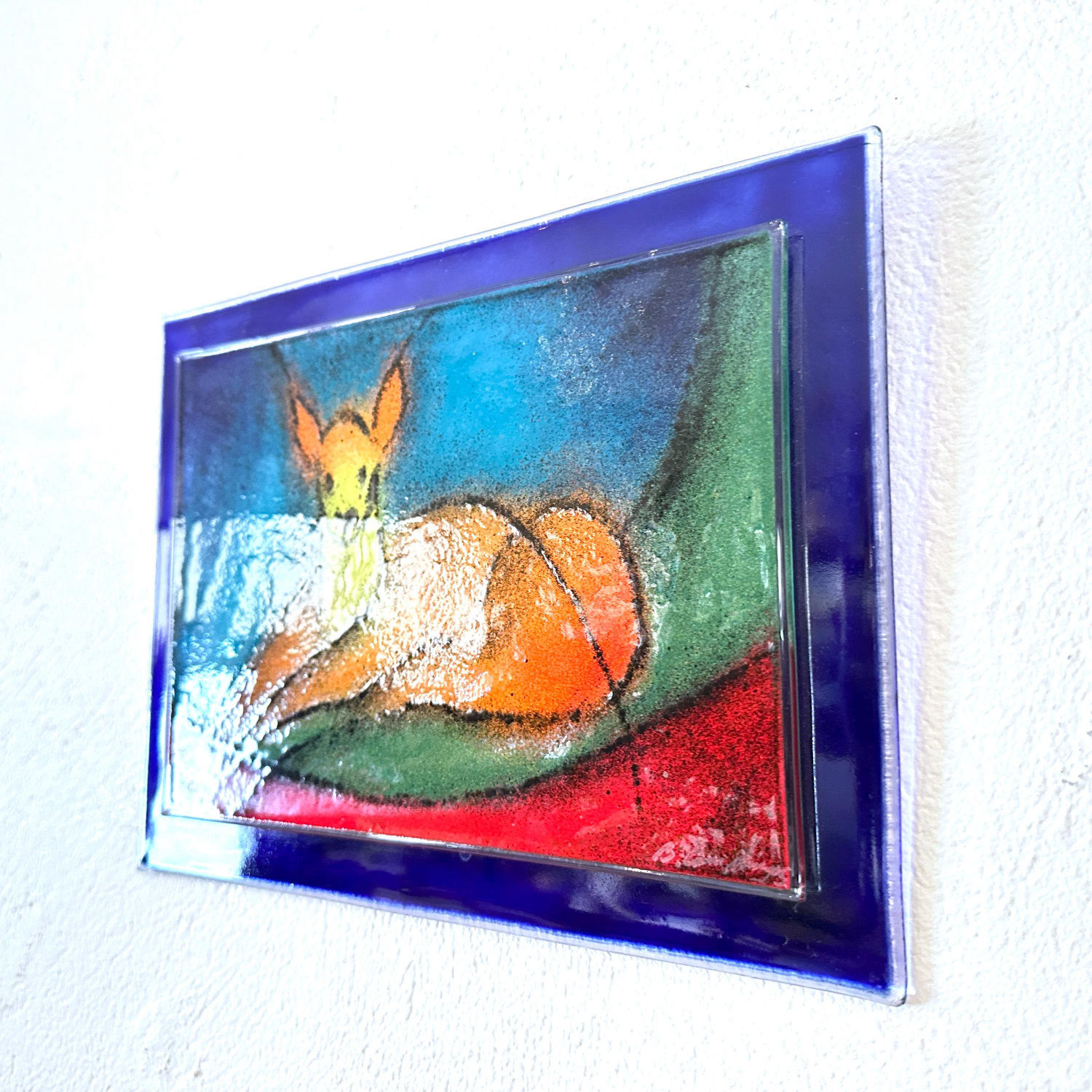 Angled view of framed glass deer panel mounted on white wall, showing shadow cast by blue frame and dimensional depth of artw