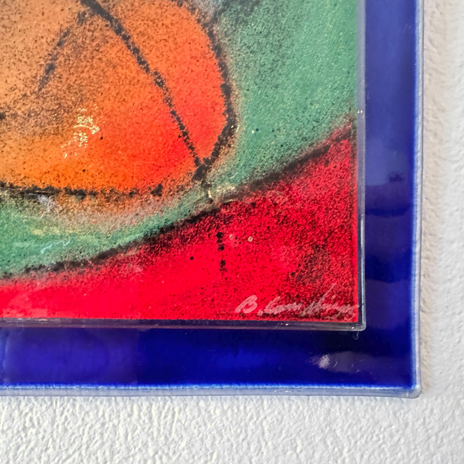 Close-up detail of glass deer artwork corner showing artist's signature, textured surface, and rich red base color with orang