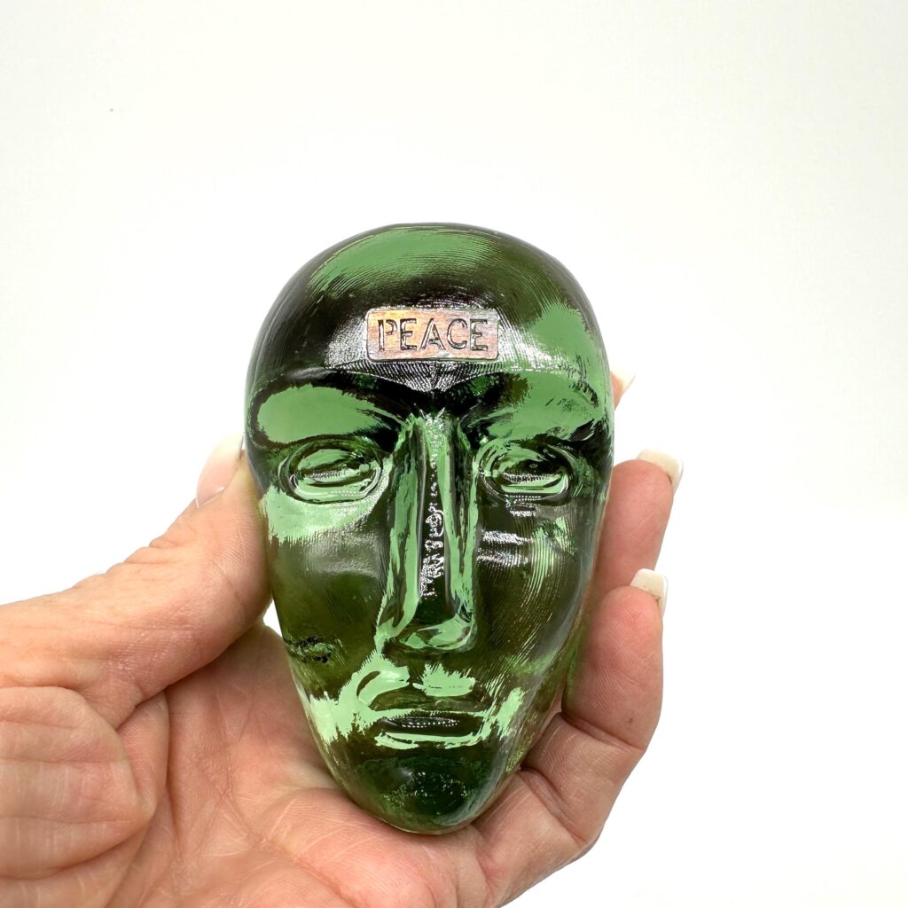 Small carved green glass head sculpture with simplified facial features, held in palm of hand for scale.