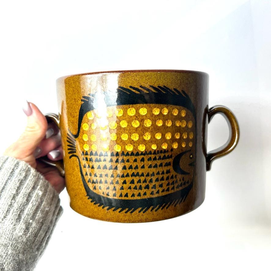 Scandinavian ceramic cache pot with stylized fish motif in ochre, brown, and rust glaze, twin handles, held in hand against w
