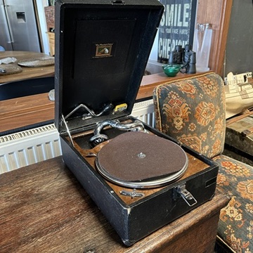 Black portable gramophone with open lid revealing tonearm and turntable, displayed on wooden shelf with decorative patterned