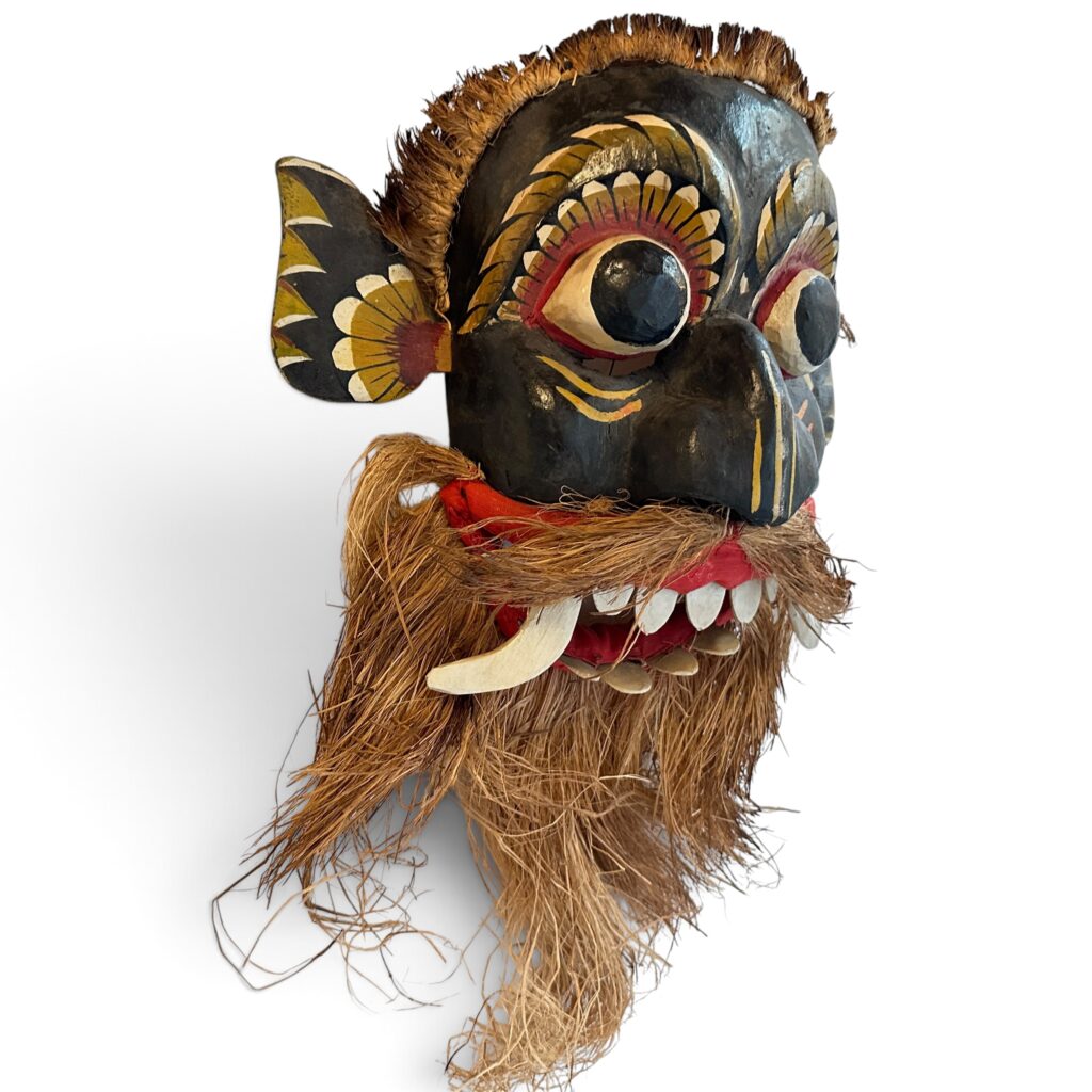 Hand-carved wooden Balinese Barong mask with protruding eyes, curved tusks, prominent teeth, and natural fiber hair